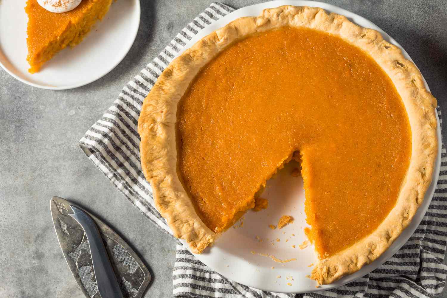Overhead view of a white pie plate of homemade sweet potato thanksgiving pie with a slice removed next to a pie server and resting on a striped napkin next to a small plate with one slice of pie