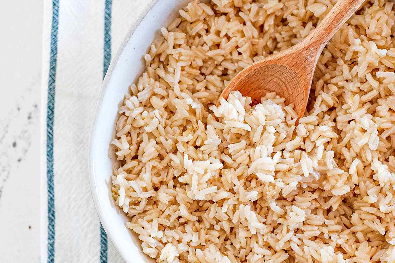 Brown rice cooked on a stove top in a white bowl with a wooden spoon