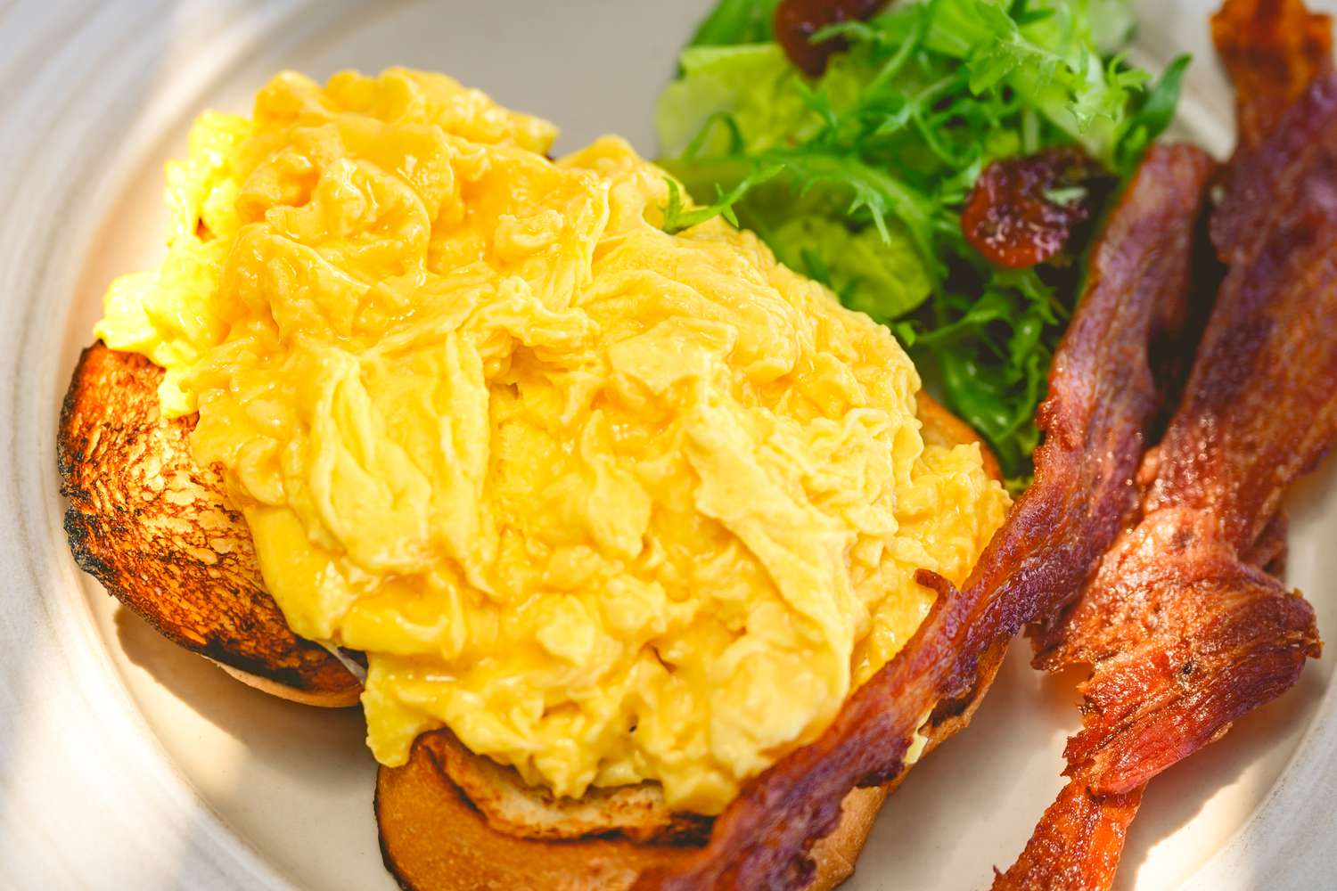Overhead view of a plate of scrambled eggs, strips of bacon, toast and greens