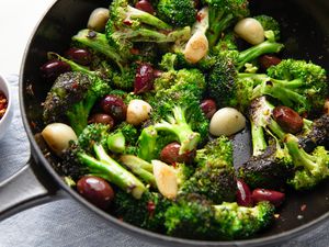 Angled view of a cast iron skillet of seared broccoli with spices, garlic cloves and olives next to a small bowl of spices