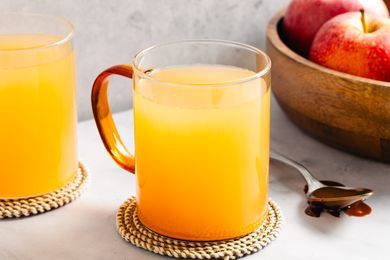 Two mugs of apple cider on coasters a wooden bowl with apples and a spoon with caramel nearby