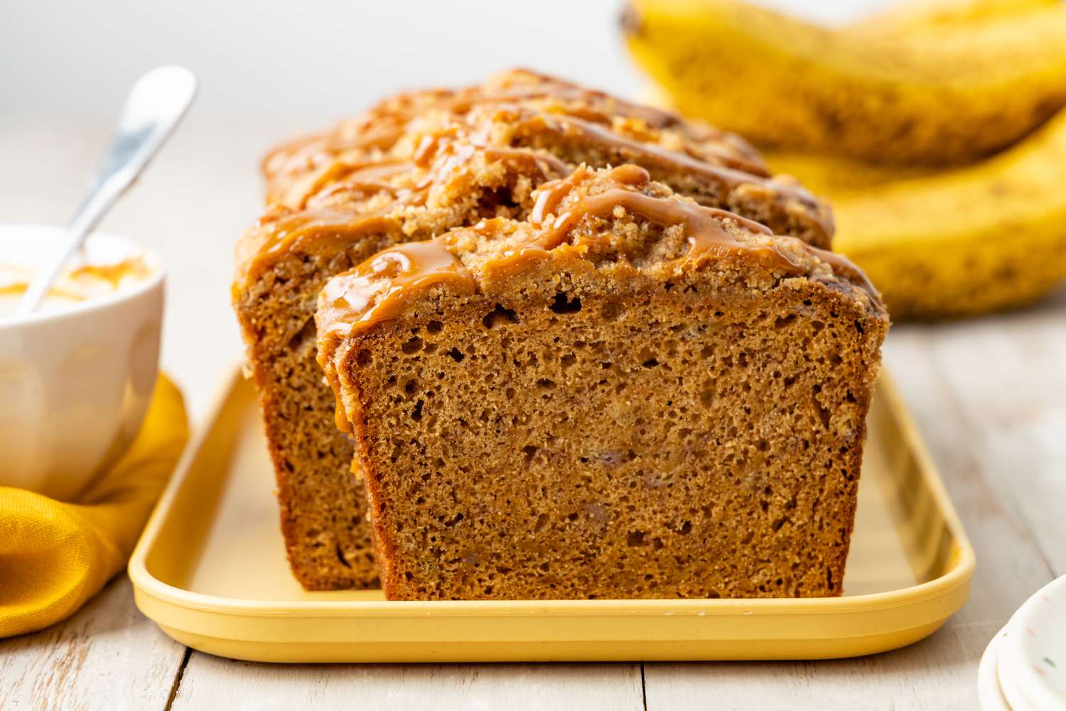 Spiced Dulce de Leche Banana Bread (Some of It Sliced) on a Platter, and in the Surroundings, a Bowl With More Dulce de Leche and Bananas
