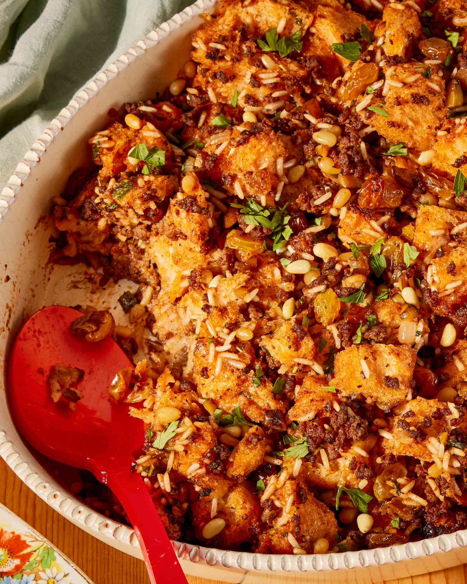 A closeup of Greek-style stuffing in an oval white serving dish, with a red serving spoon in the right corner where some of the stuffing has been scooped out