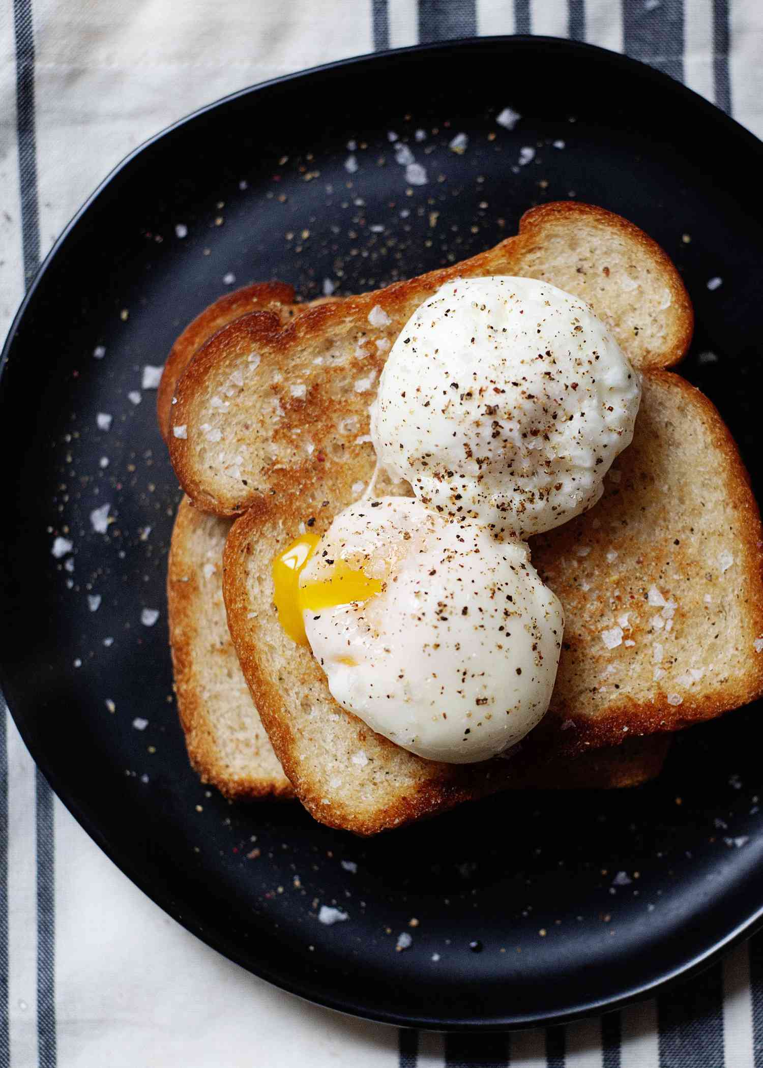 Two poached eggs on top of two pieces of stacked toast. One egg has a slightly runny yolk. Salt and pepper are sprinkled on the eggs, toast and black plate.
