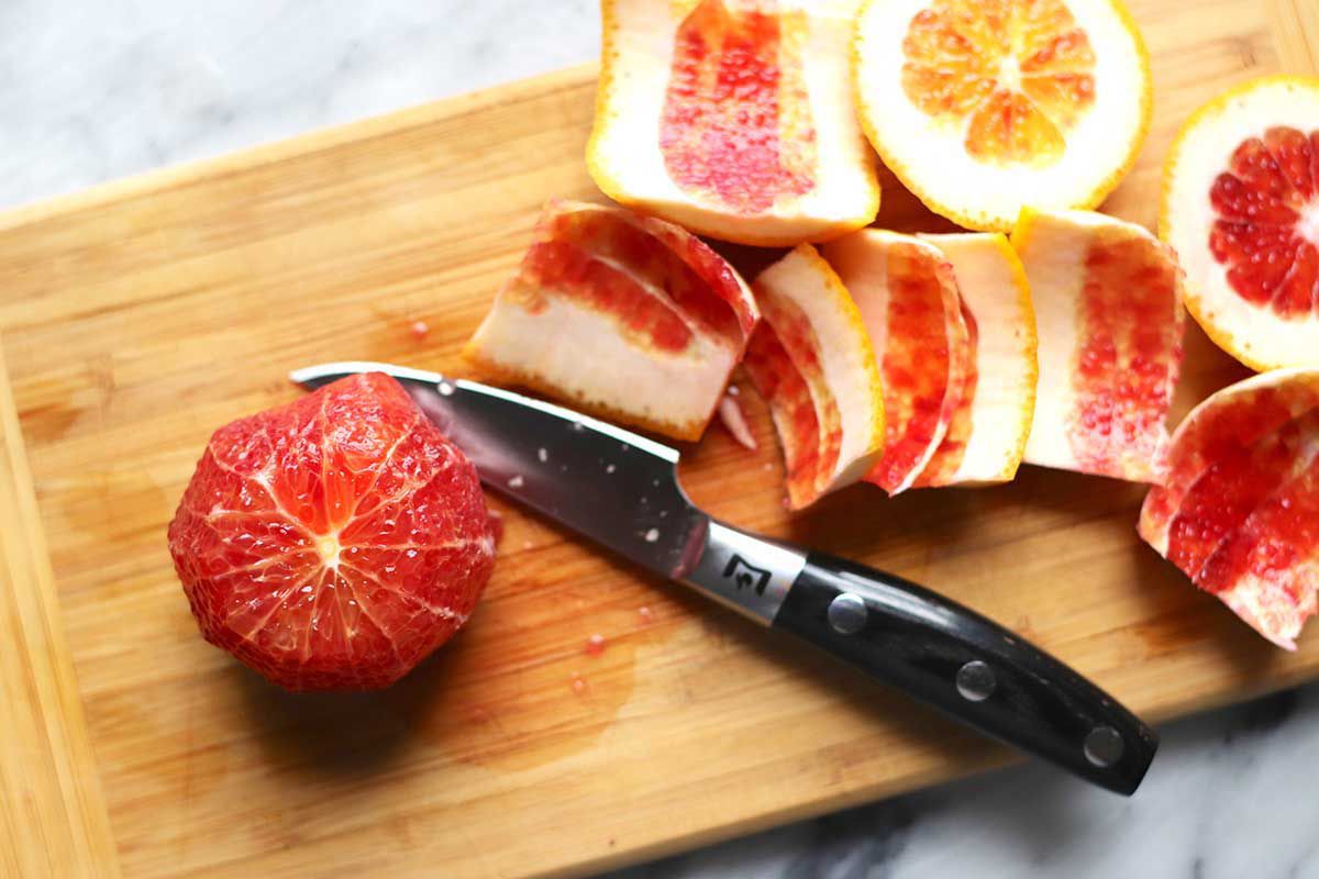 An overhead view of a wooden cutting board with a paring knife set next to a blood orange with the skin removed. The peels are on the right of the knife.