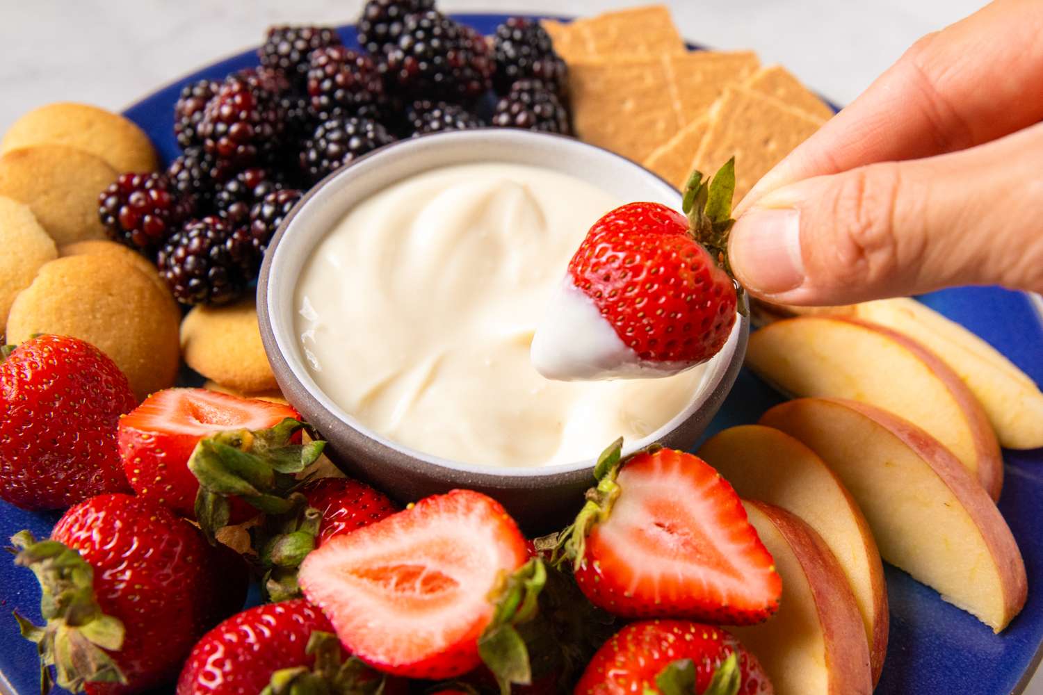 Creamy dip surrounded by strawberries blackberries apple slices vanilla wafers and graham crackers with a strawberry being dipped by a hand