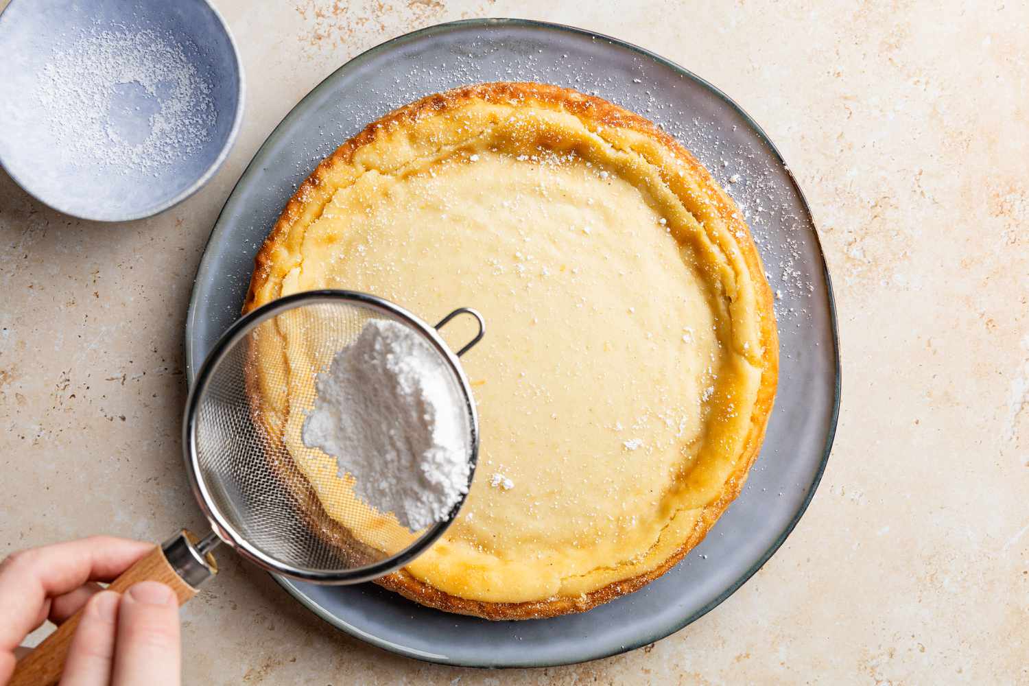 cake on a plate, hand sifting powdered sugar around the edges for Migliaccio (Italian Ricotta and Semolina Cake)
