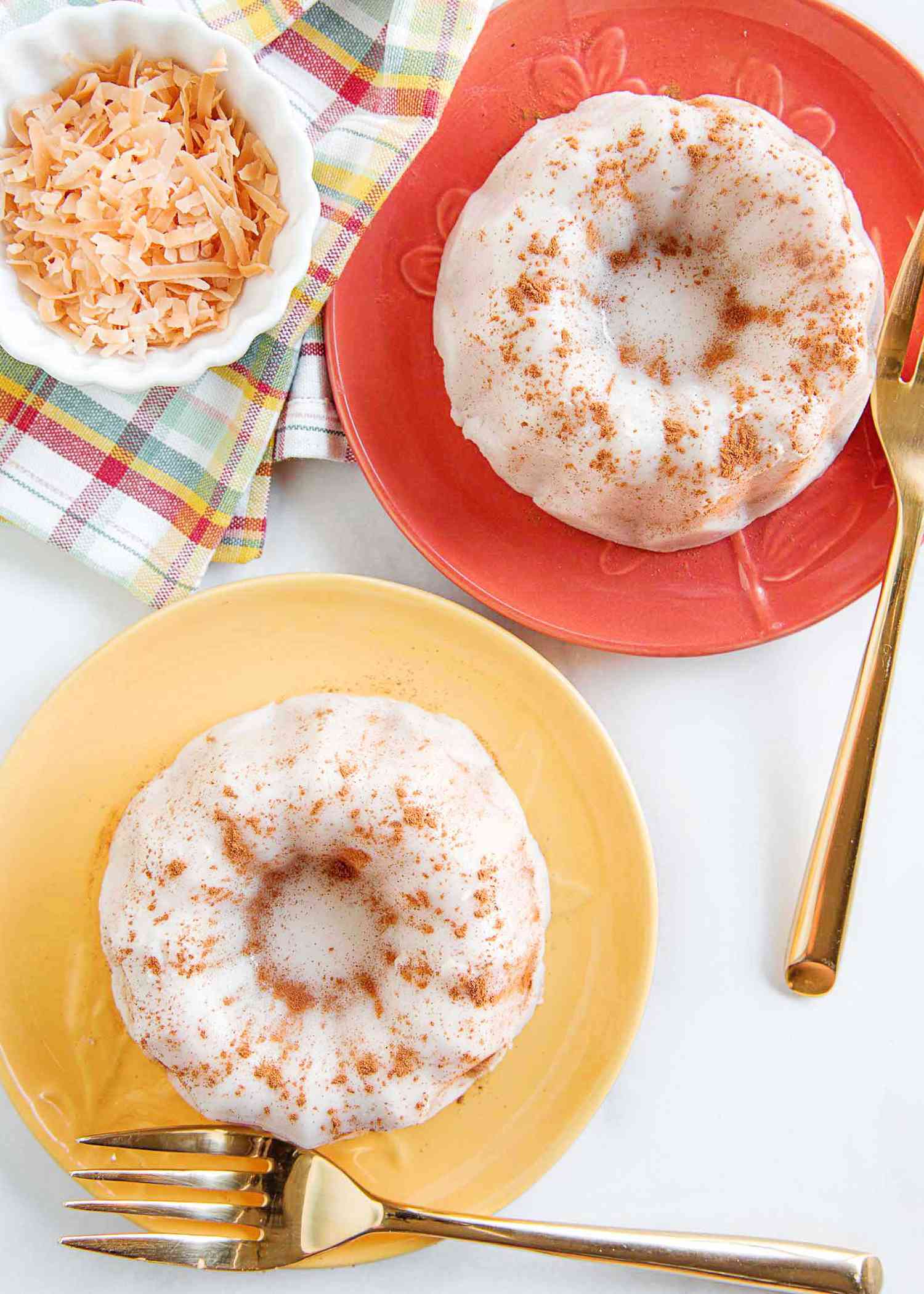 Vertical photo of two plates with coconut pudding and gold forks. The coconut pudding is coated in ground cinnamon. The lower plate is light yellow and the plate in the upper right is coral. A small container of toasted shredded coconut is to the left of the coral plate. A striped dish cloth is underneath the coconut.