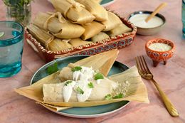 A Plate of Chicken Tamales with Chile Verde Topped with Crema, Cotija Cheese, and Some Cilantro at a Table Setting with a Platter of More Tamales, Utensils, a Small Bowl of Cotija Cheese, a Small Bowl of Crema, a Glass of Water, and a Small Vase