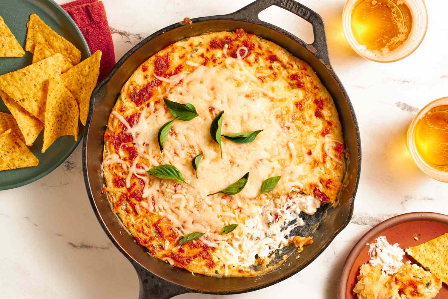 Baked dip with cheese and basil in a castiron skillet chips and drinks served around it