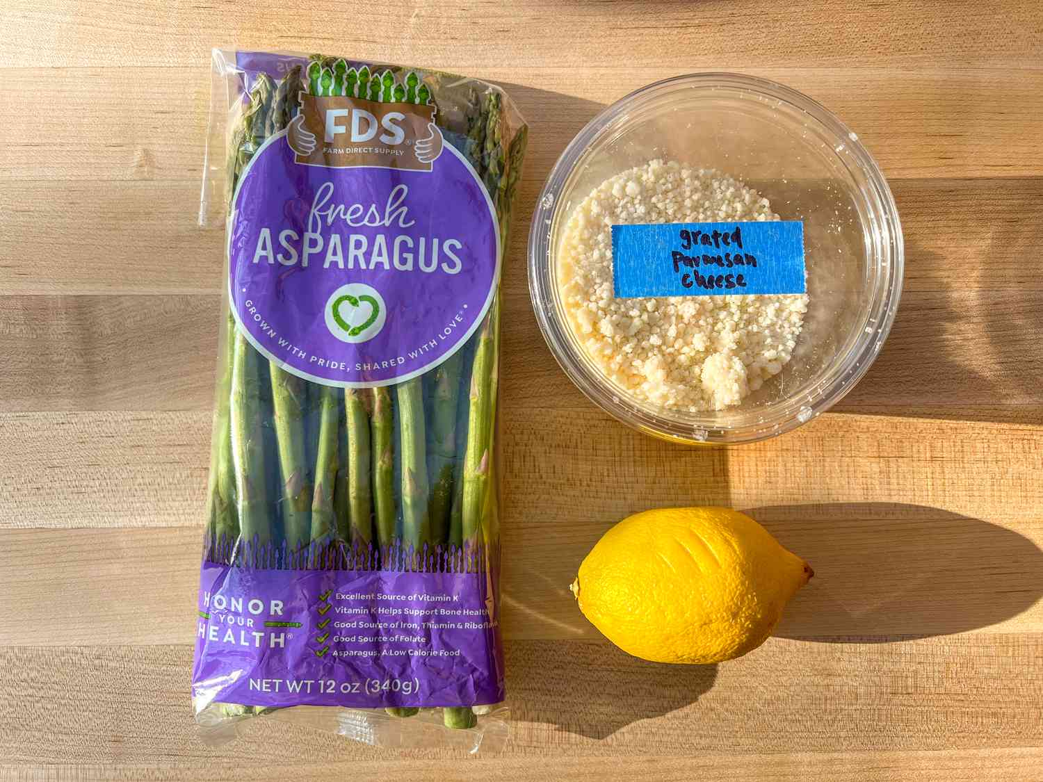 Packaged asparagus, grated cheese in a container, and a lemon arranged on a wooden surface