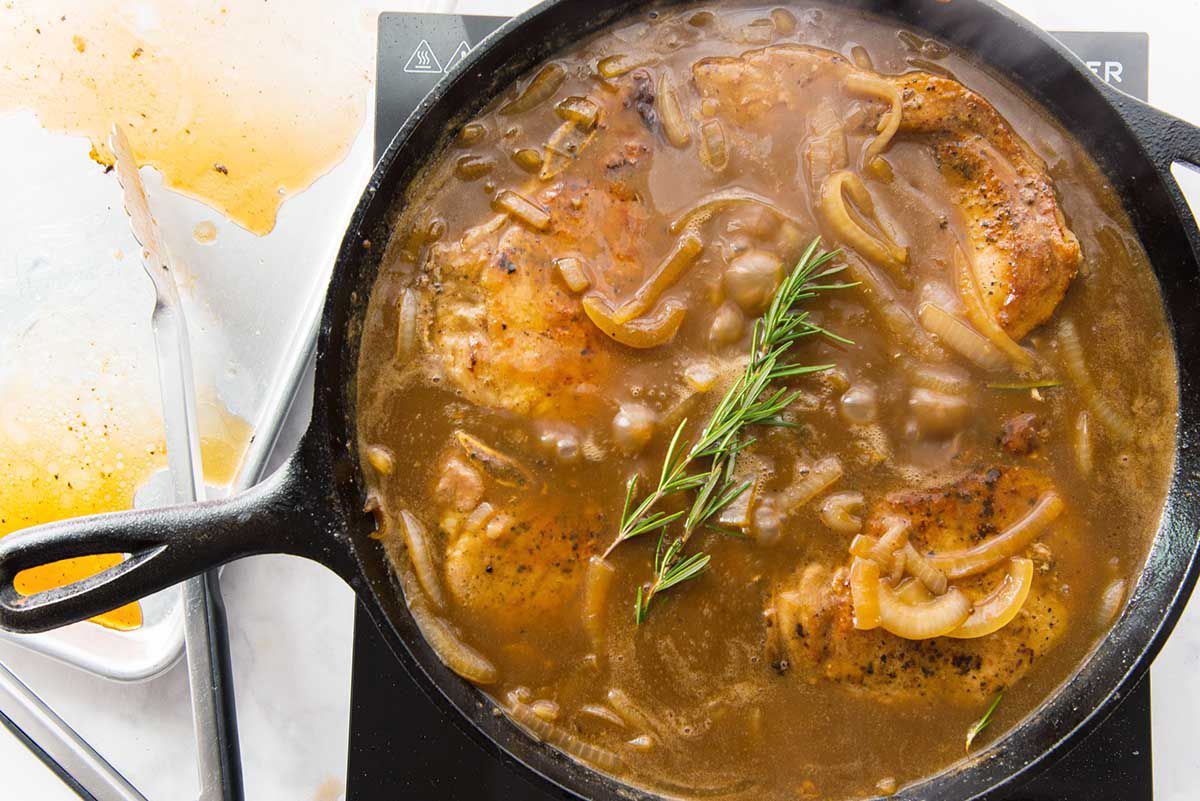 Onions, pork chops and a sprig of rosemary in a cast iron skillet filled with gravy and being stirred with a wooden spoon.