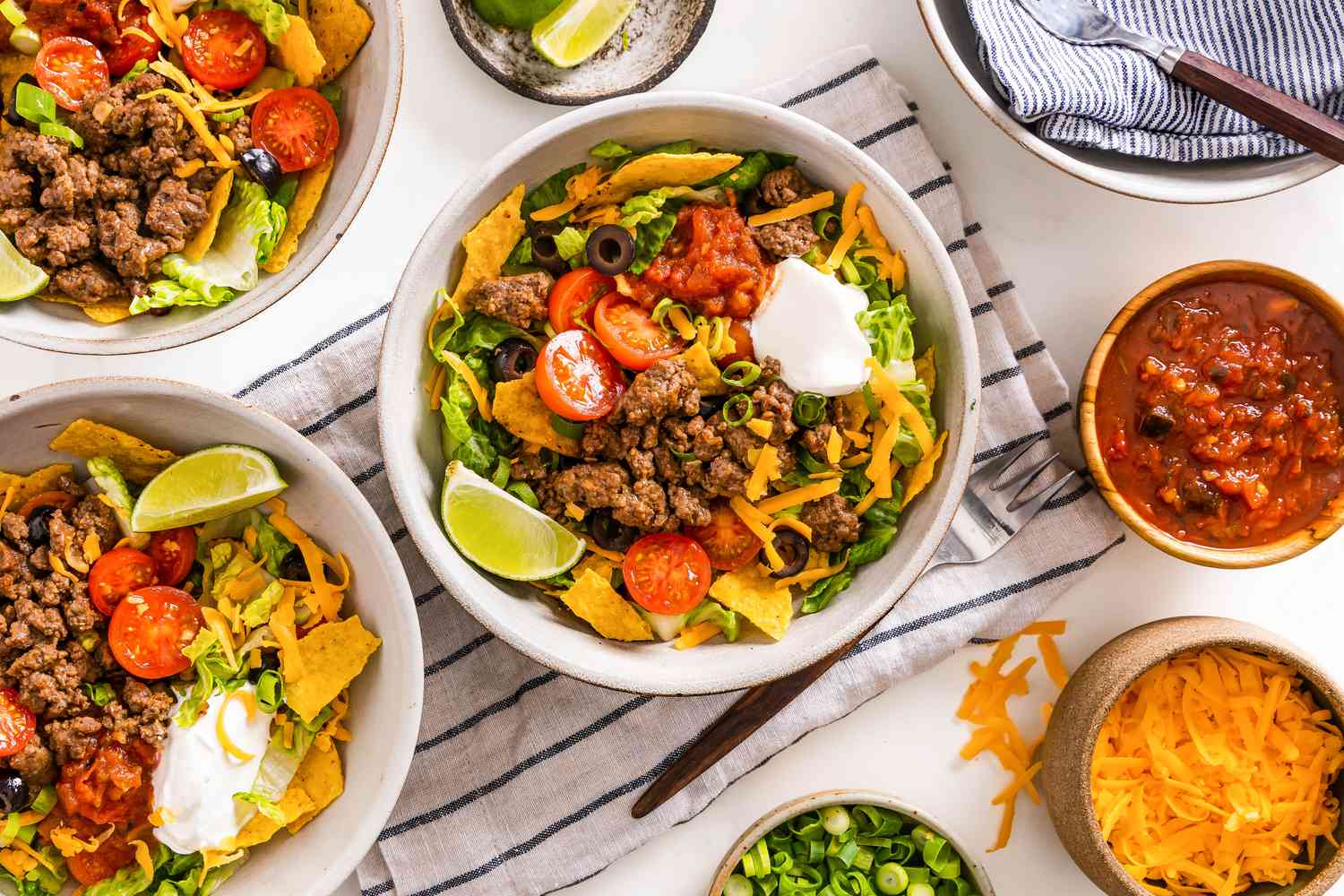 Bowl of Taco Salad Surrounded by Two More Bowls With Servings of Taco Salad, a Bowl of Salsa, a Bowl of Quartered Limes, a Bowl of Cheese, a Bowl of Sliced Green Onions, and a Bowl With Utensils, All on a Kitchen Towel