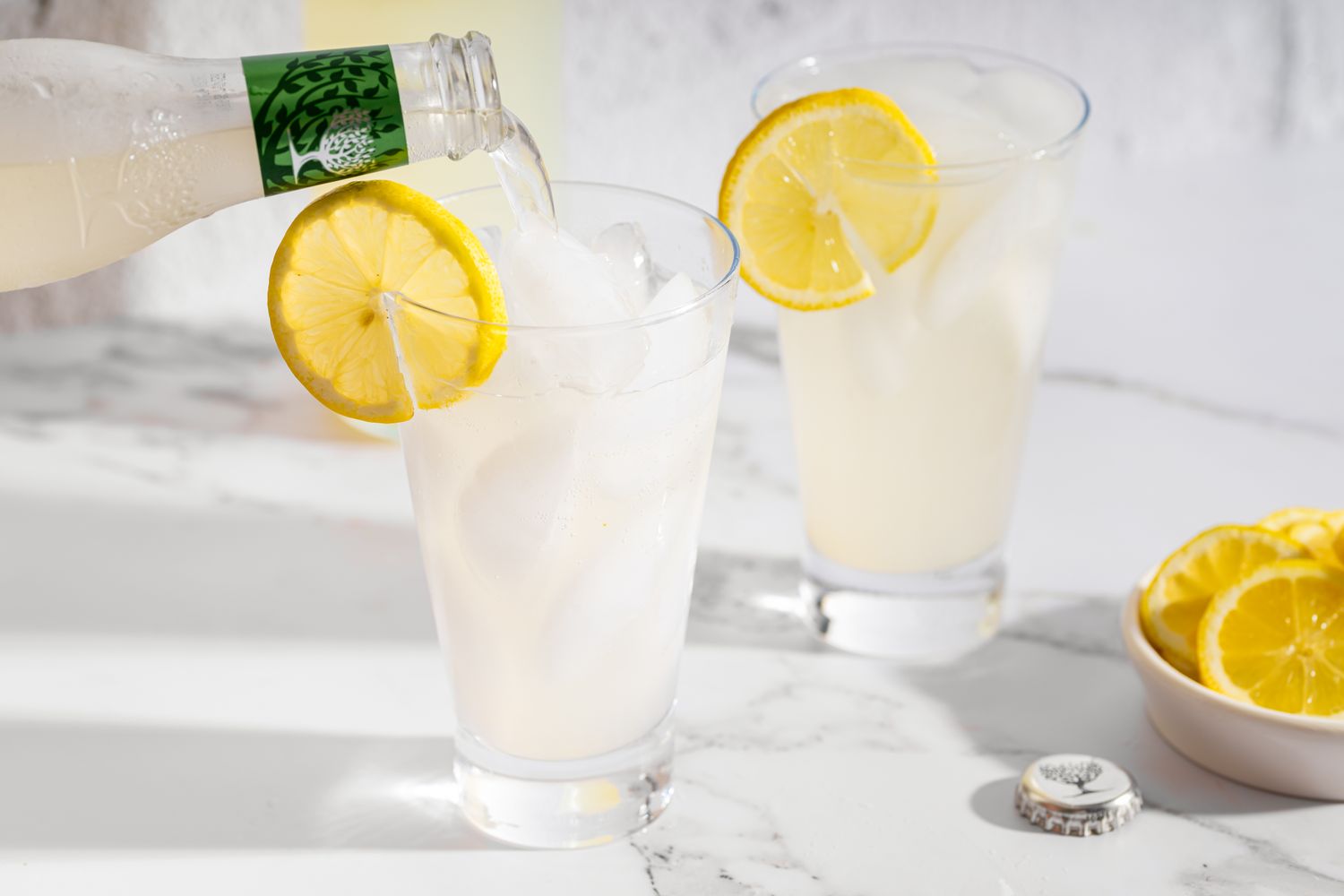 Two glasses of ginger lemonade one being filled garnished with lemon slices on a marble surface next to a small bowl of lemon slices