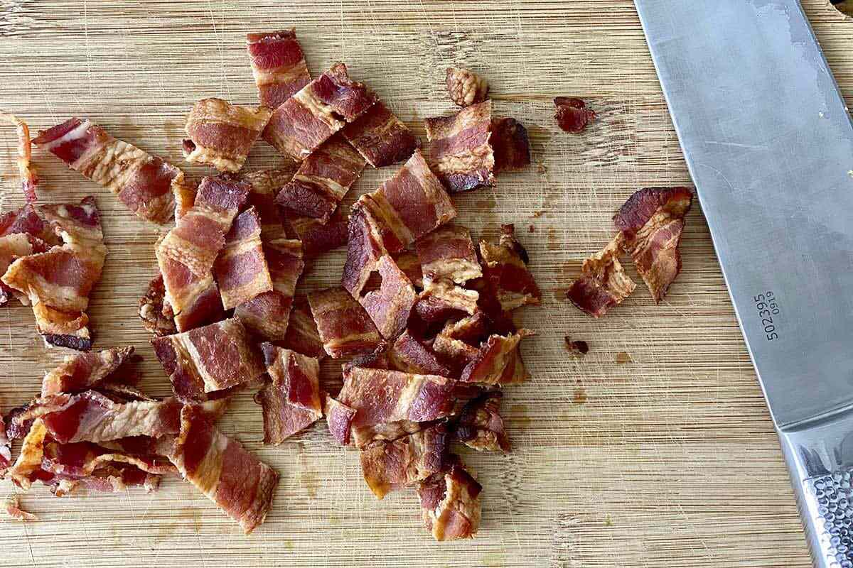 Chopped, cooked bacon on a cutting board with a chef's knife for Kale and Brussels Sprout Salad with Dried Cranberries and Maple Dijon Vinaigrette.