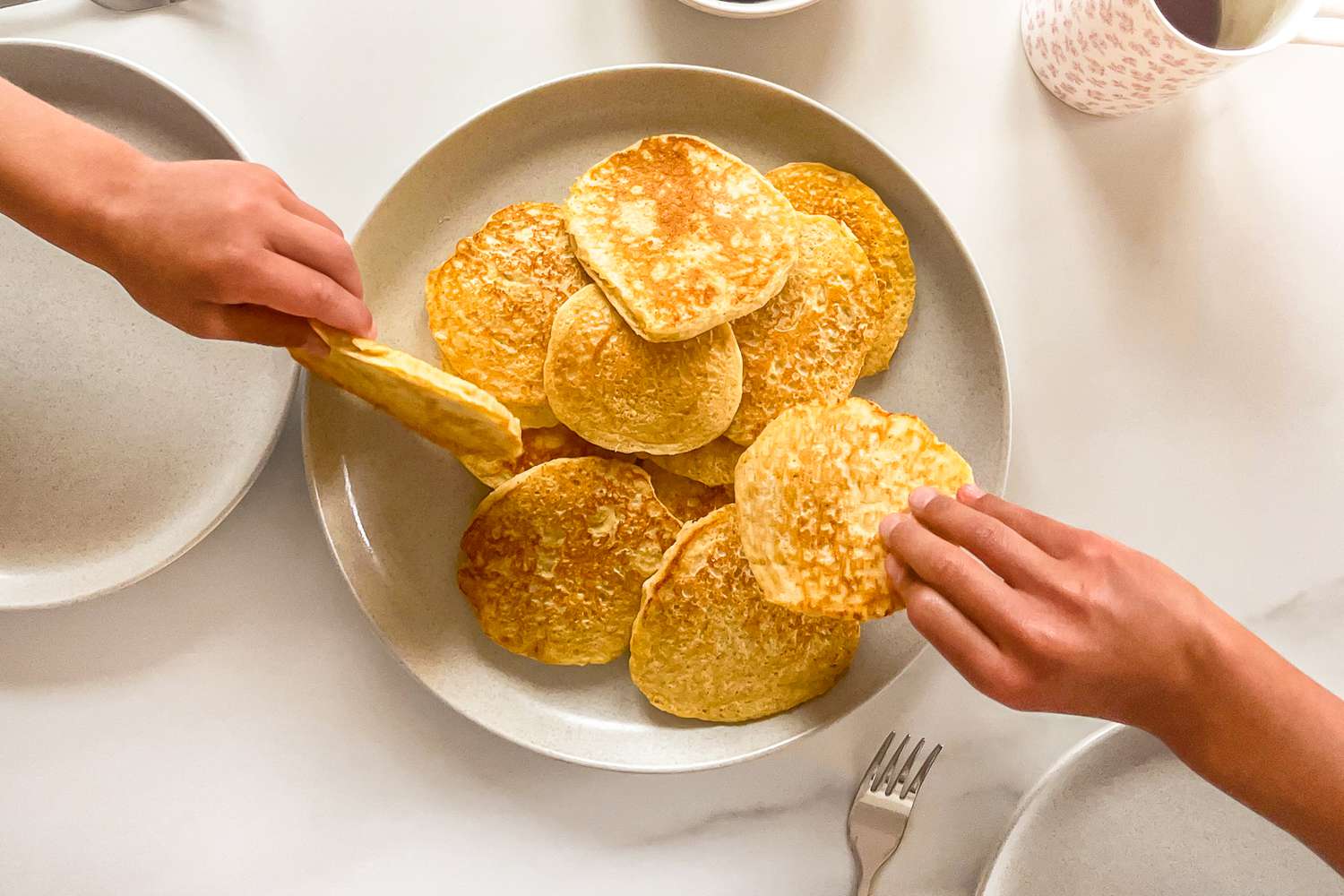 Two hands taking pancakes from a plate on a table with other tableware