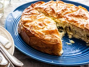 Side view of a flaky spring pie on a large blue platter. One slice of the pie is missing and the cheesy artichoke filling is visible on the plate. The lower left has a pile of forks on a white plate. A grey linen tablecloth is on the table and a glass is in the upper left corner.