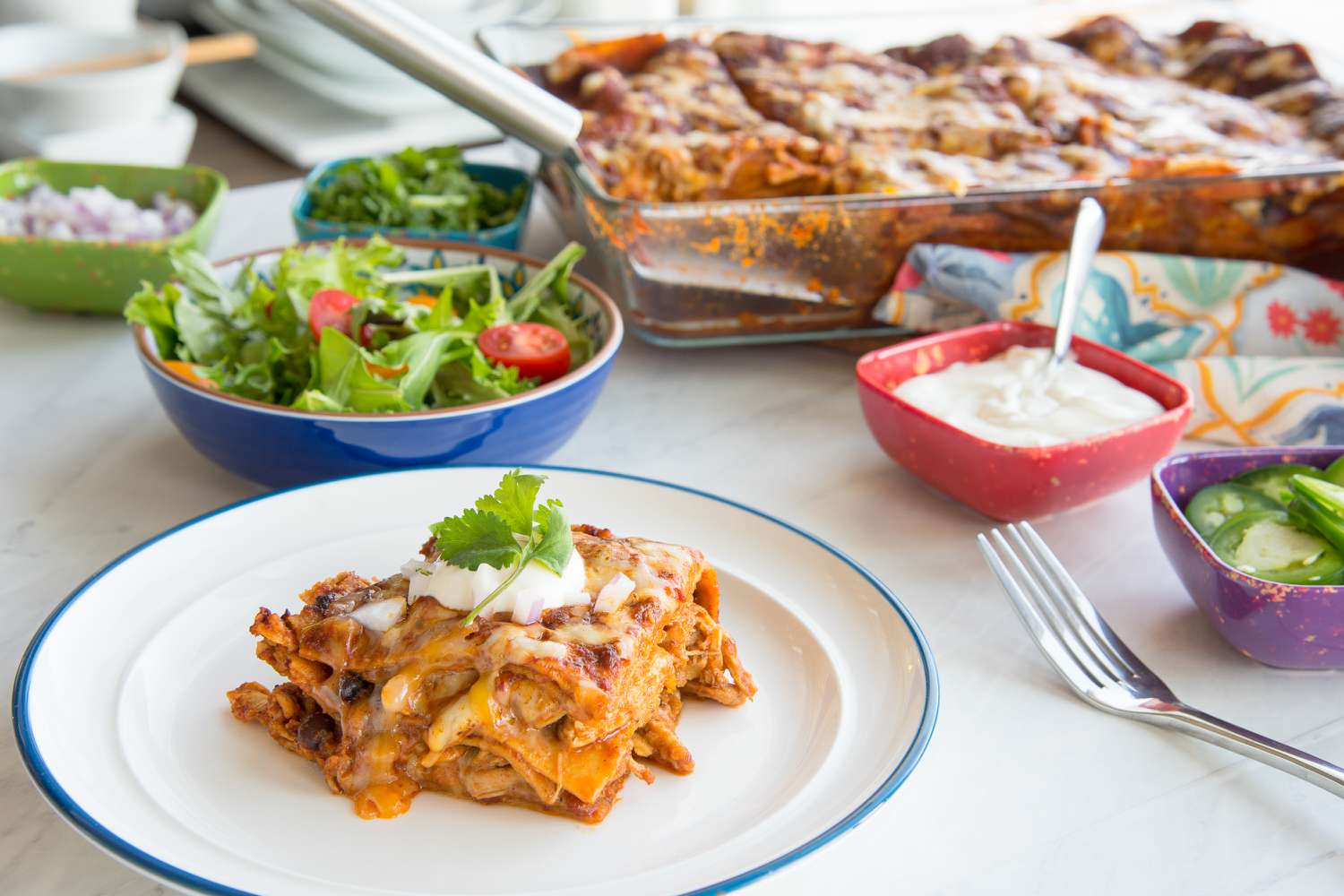 Serving of Easy Chicken Enchilada Casserole on a Plate Topped with a Dollop of Sour Cream, Chopped Onions, and Cilantro, and in the Background, a Bowl with Side Salad, Smaller Bowls with More Toppings, and a Casserole Dish with More Casserole