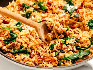 Angled view of a wooden spoon in a skillet of orzo, italian sausage, sun-dried tomatoes and baby spinach