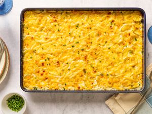 Overhead view of a 9x13 sheet pan of Sheet Pan Au Gratin Potatoes recipe on a countertop with forks, a serving spoon, glasses and a small bowl of chives