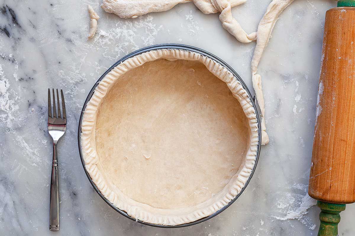 A springform pan with uncooked quiche crust inside. The crust is decorated with fork tine lines around the outside. A wooden rolling pin and scraps of dough are to the right of the pan. A fork is to the left. All are on a marble countertop.