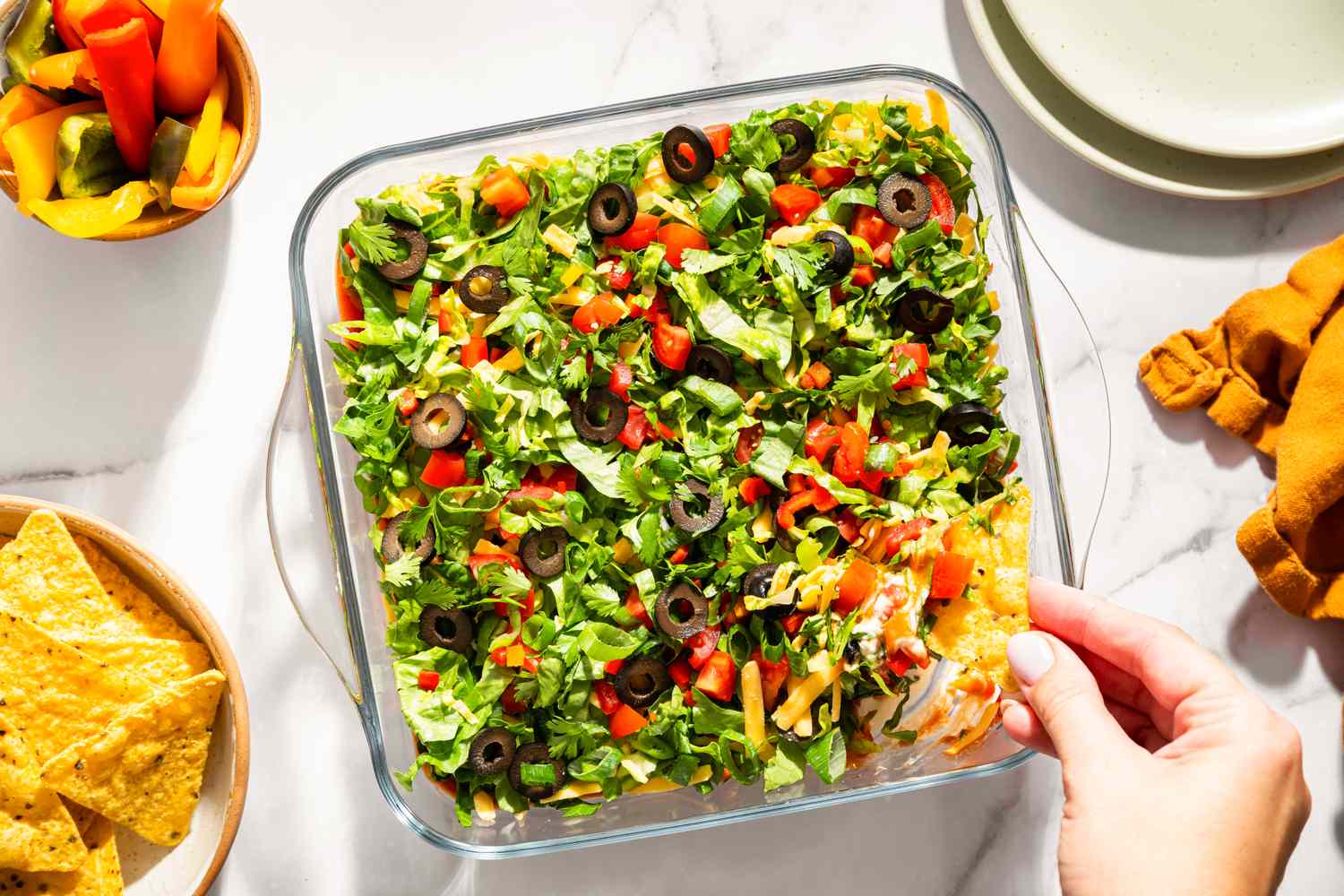 Layered nacho dip with vegetables and olives in a glass dish surrounded by chips and a hand holding a serving