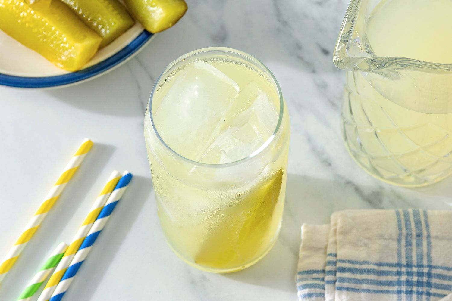 Pickle Lemonade in a glass with a straw, plate of pickle spears, extra straws, and a pitcher of lemonade