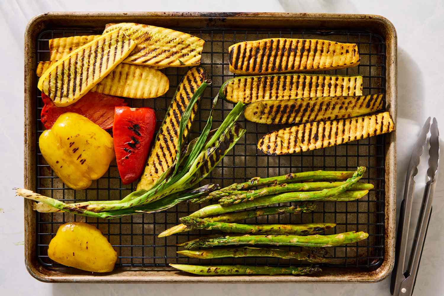 Grilled vegetables including bell peppers, zucchini, and asparagus on a baking sheet with tongs to the side