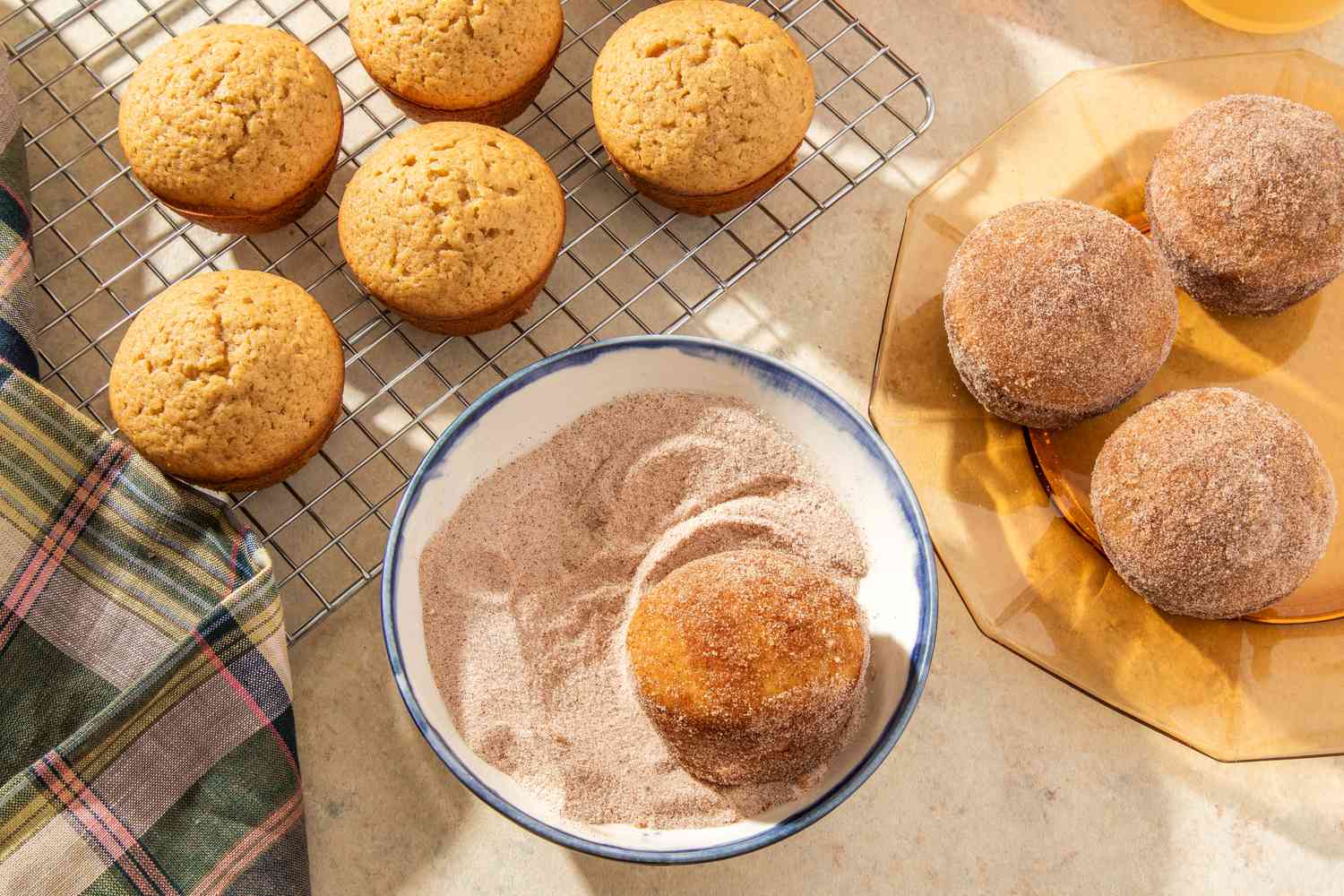 muffins on a cooling rack with a muffin in a bowl of cinnamon sugar and finished muffins on a plate