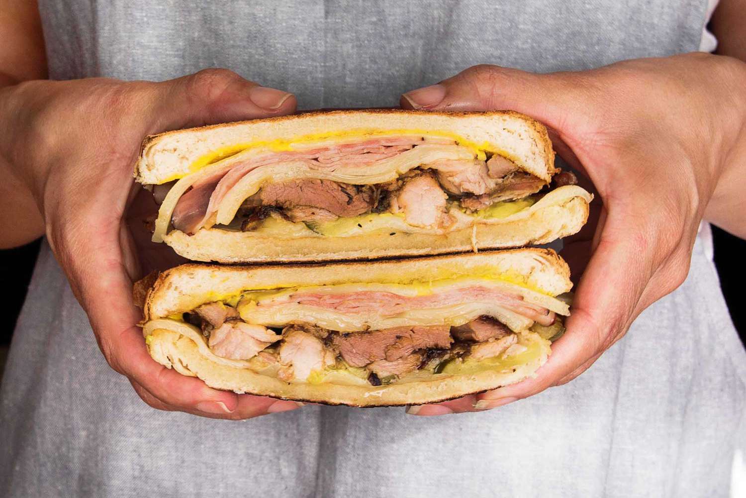 A person is holding two halves of a cuban sandwich. The person is wearing a grey apron an the shredded pork, sliced ham, sliced cheese, mustard and mayonnaise are visible.