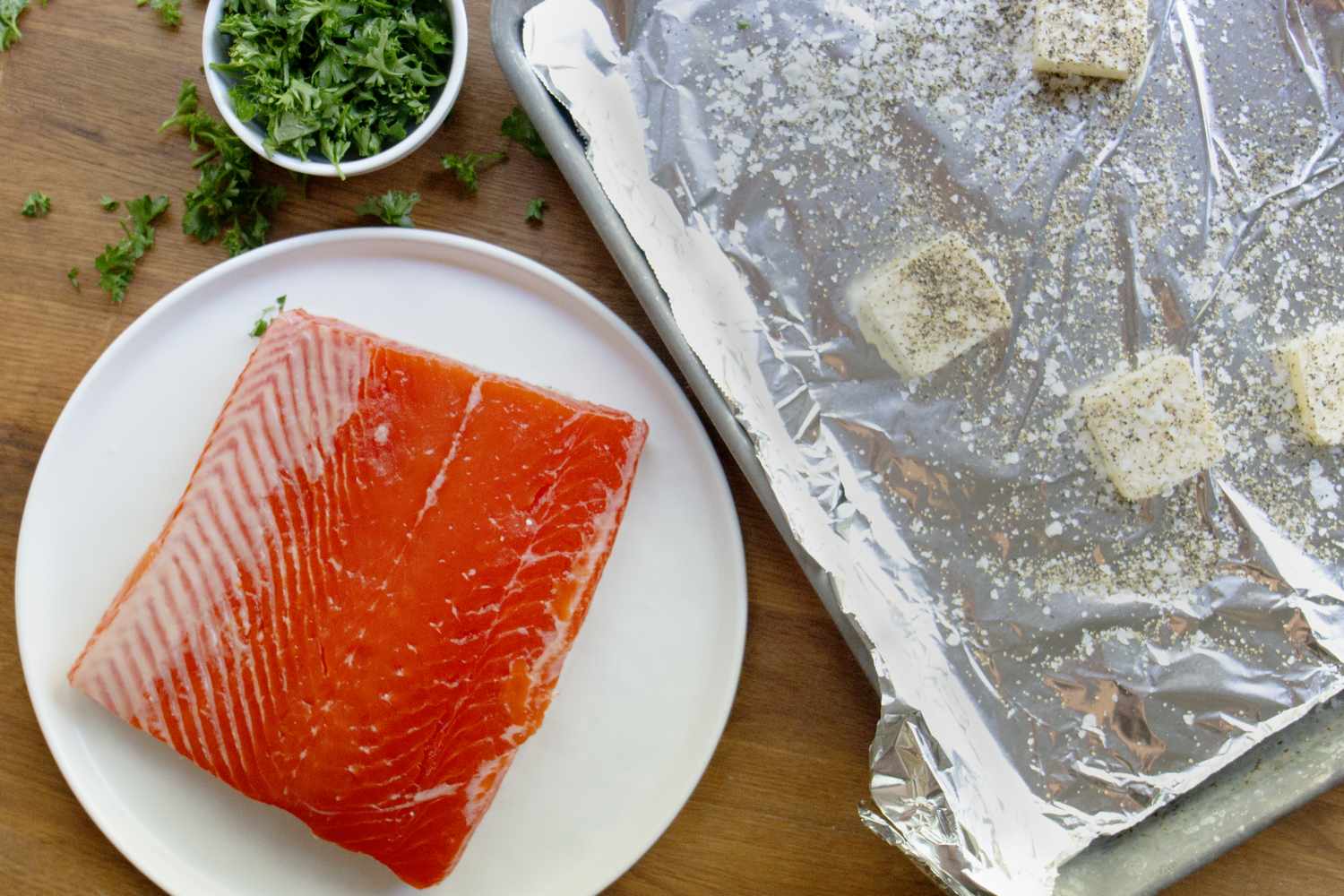 Raw salmon filet on a plate next to a prepared baking sheet with seasoned butter cubes and a bowl of chopped parsley