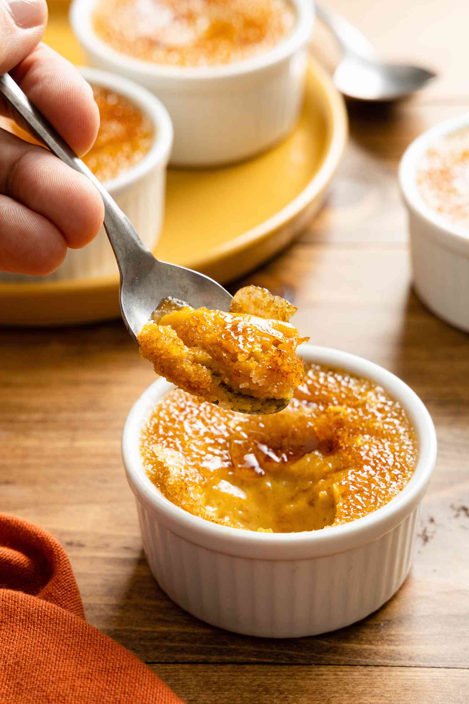 3-Ingredient Pumpkin Spice Crème Brûlée on a Spoon and More in Ramekin Below It, Surrounded by More Ramekins, Another Spoon, and a Table Napkin