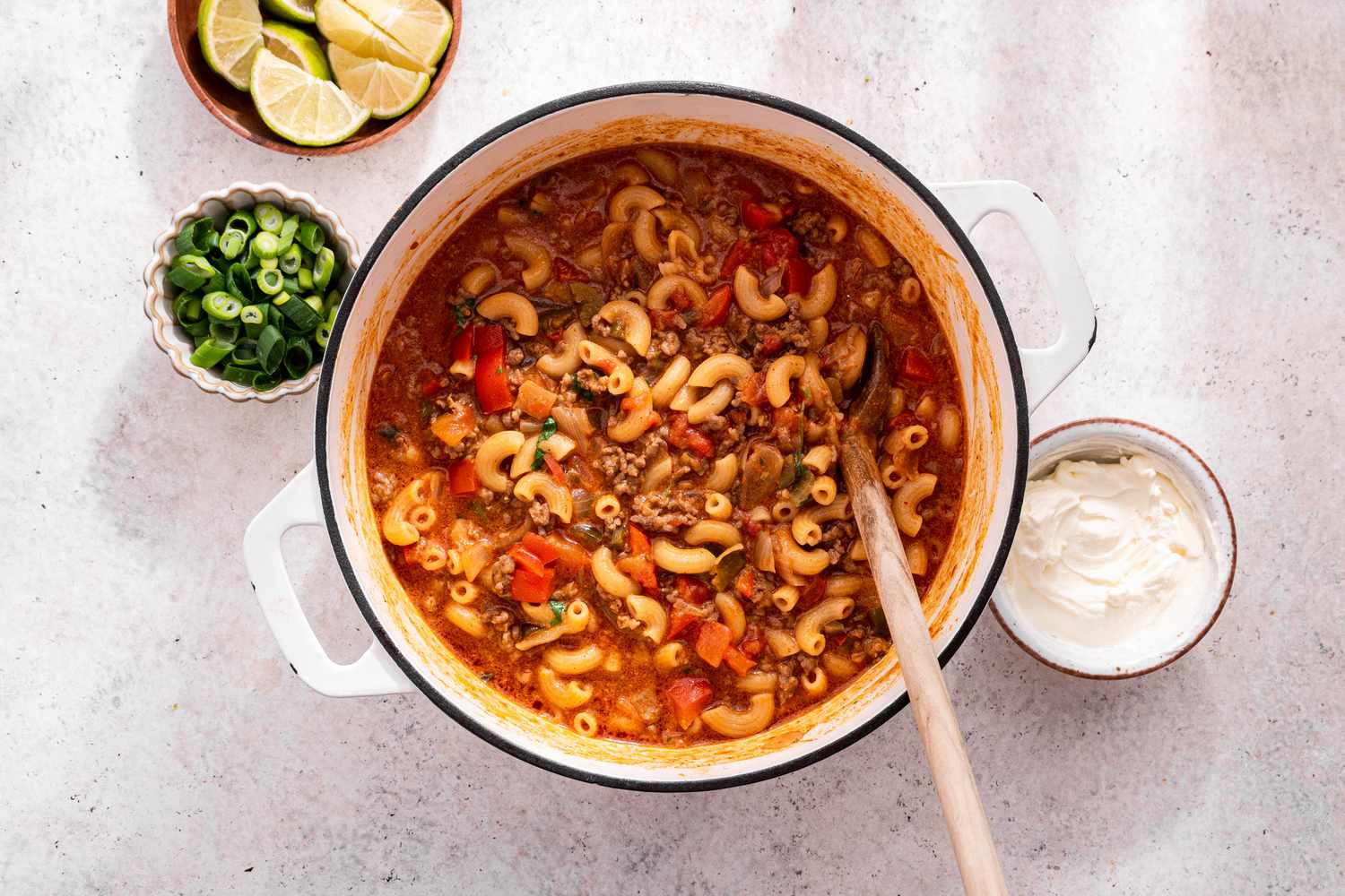 One-Pot Taco Salad in a Dutch Oven With a Wooden Spoon Surrounded by Bowls of Toppings (a Bowl of Lime Wedges, a Bowl of Sliced Scallions, and a Bowl of Sour Cream)