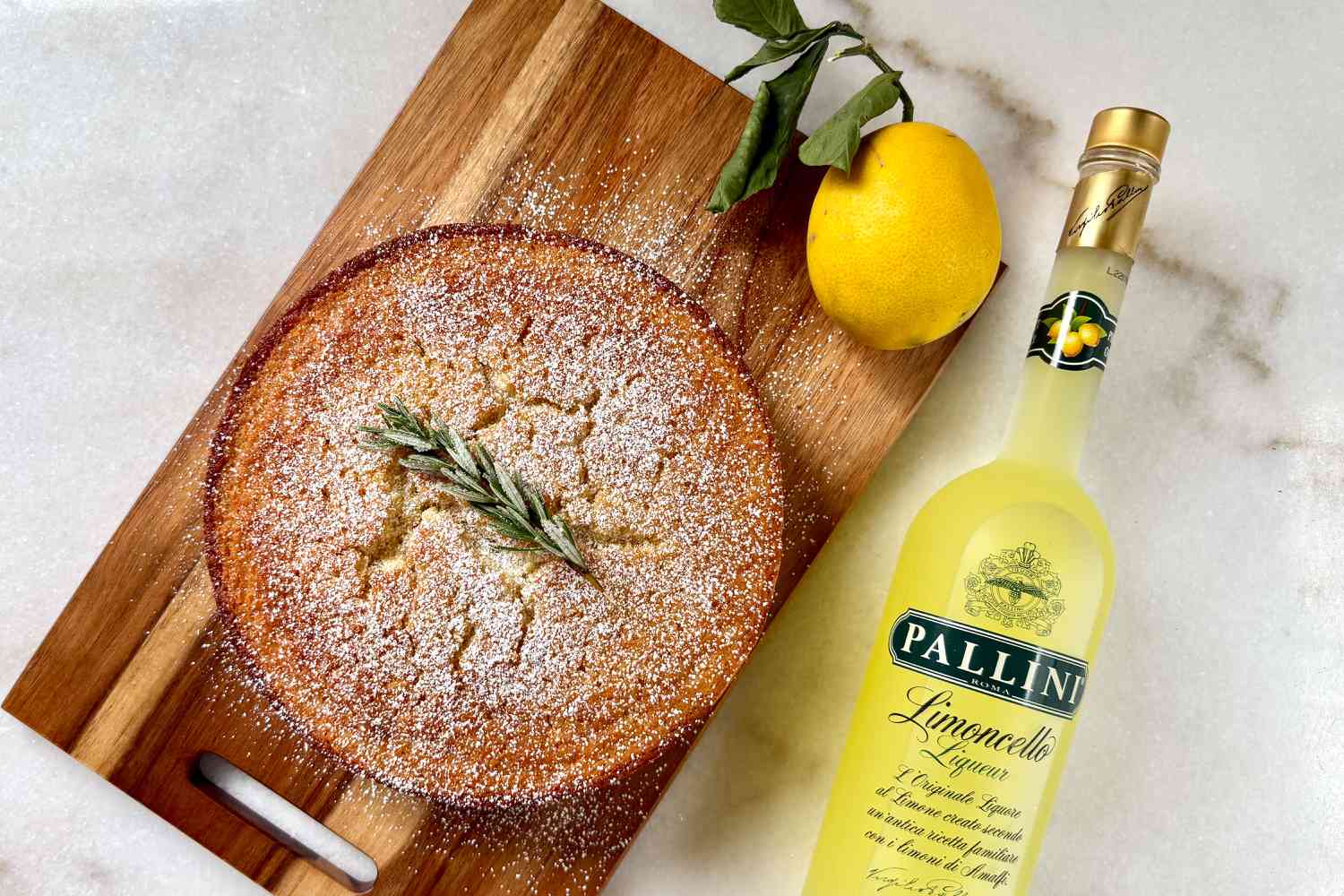Meghan Markle's Lemon Olive Oil Cake on a cutting board, sprinkled with powdered sugar and topped with a sprig of sugared rosemary, bottle of Limoncello Liquor, and a whole lemon