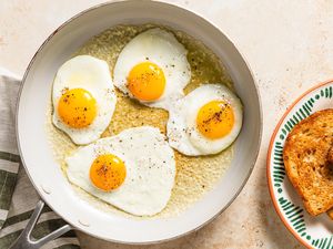 Four fried eggs in a pan next to a plate of toasted bread