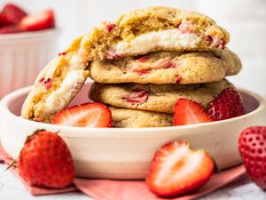Stack of Strawberry Cheesecake Cookies With the Top Cookie Cut in Half to Showcase Cheesecake Filling in a Small Plate With a Few Halved Strawberries, and in the Surroundings, a Bowl of Strawberries and Some Free Flowing Strawberries (Some Whole, Some Halved) on the Counter, All on a Pink Table Napkin 