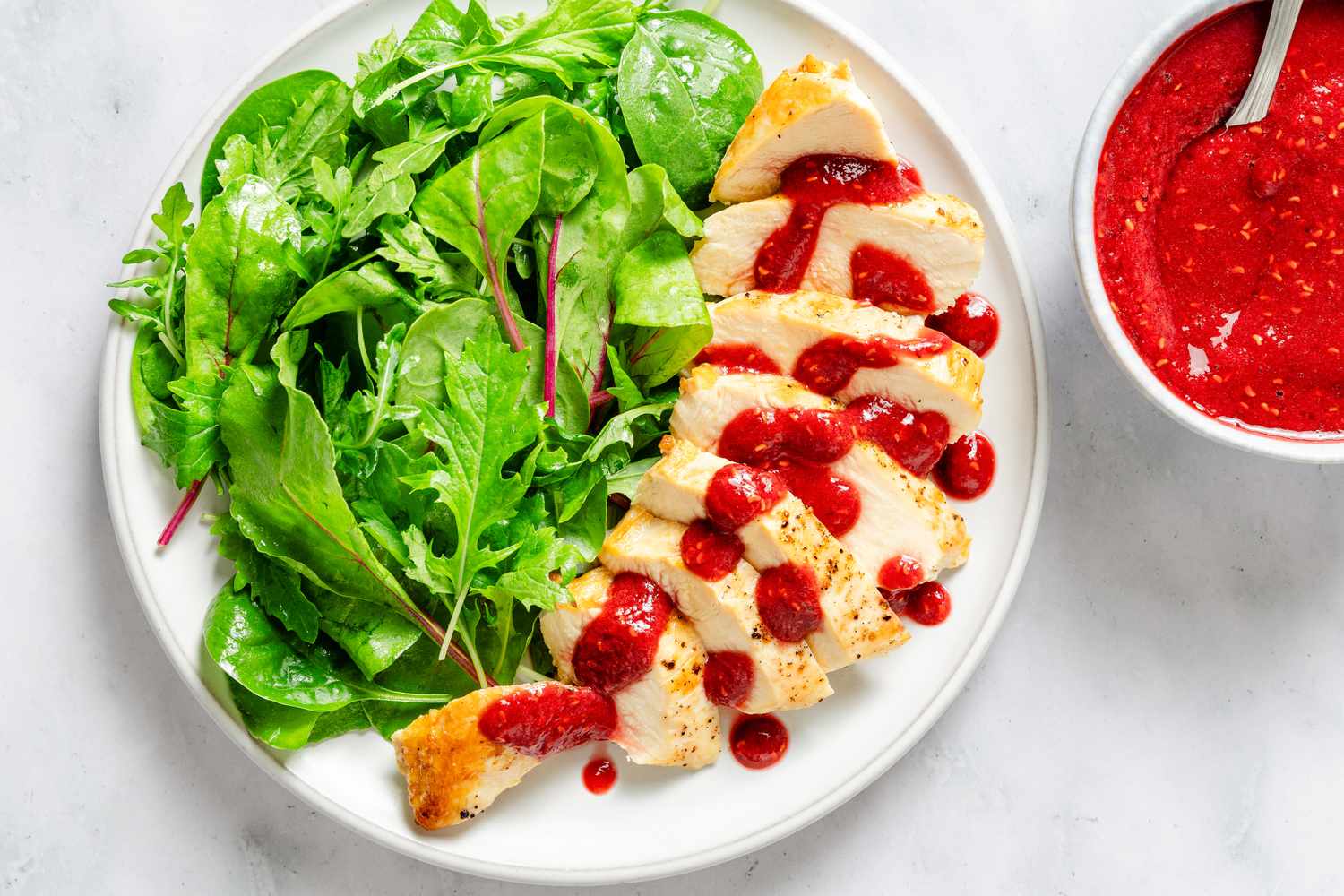 Raspberry-Soy Sauce in a small bowl with a spoon and a plate of chicken with sauce drizzled on top