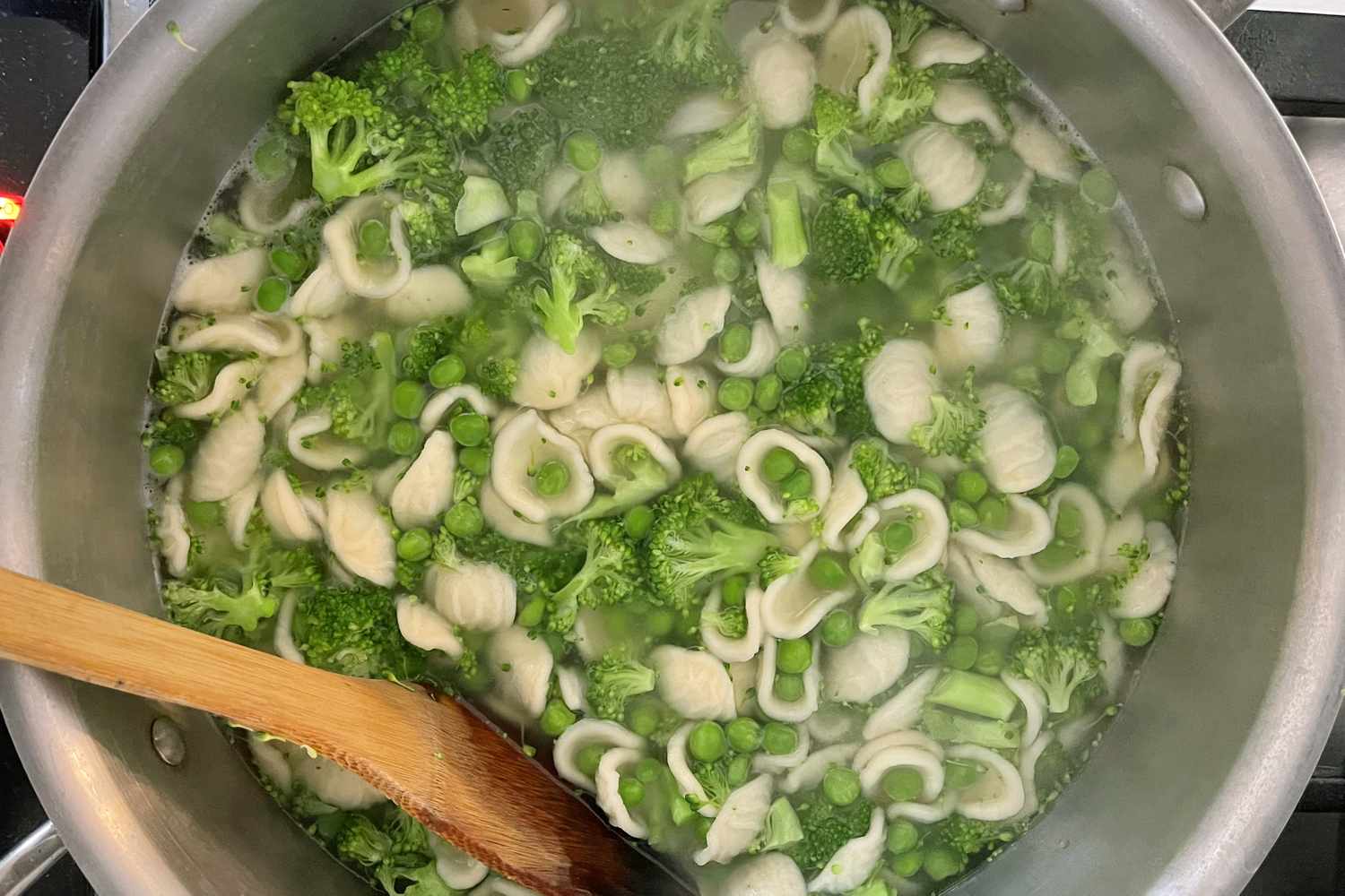 Pot containing cooked orecchiette pasta with broccoli and green peas wooden spoon stirring the mixture