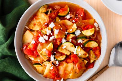 Overhead view of a white bowl of Zucchini Con Patate on a table next to a spoon and a green napkin