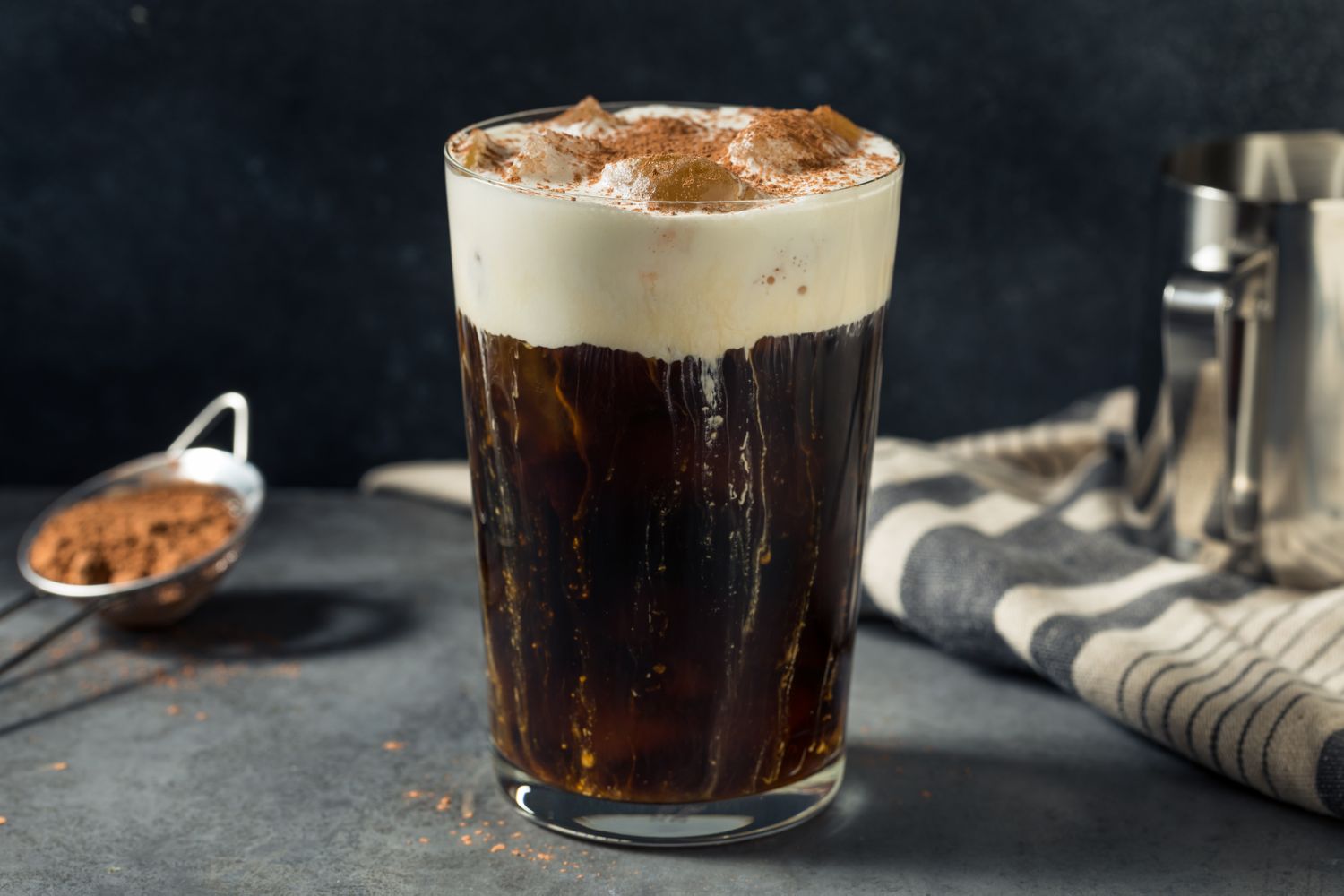 A glass of German iced coffee with cream and ice served on a gray surface with a napkin and a coffee scoop nearby