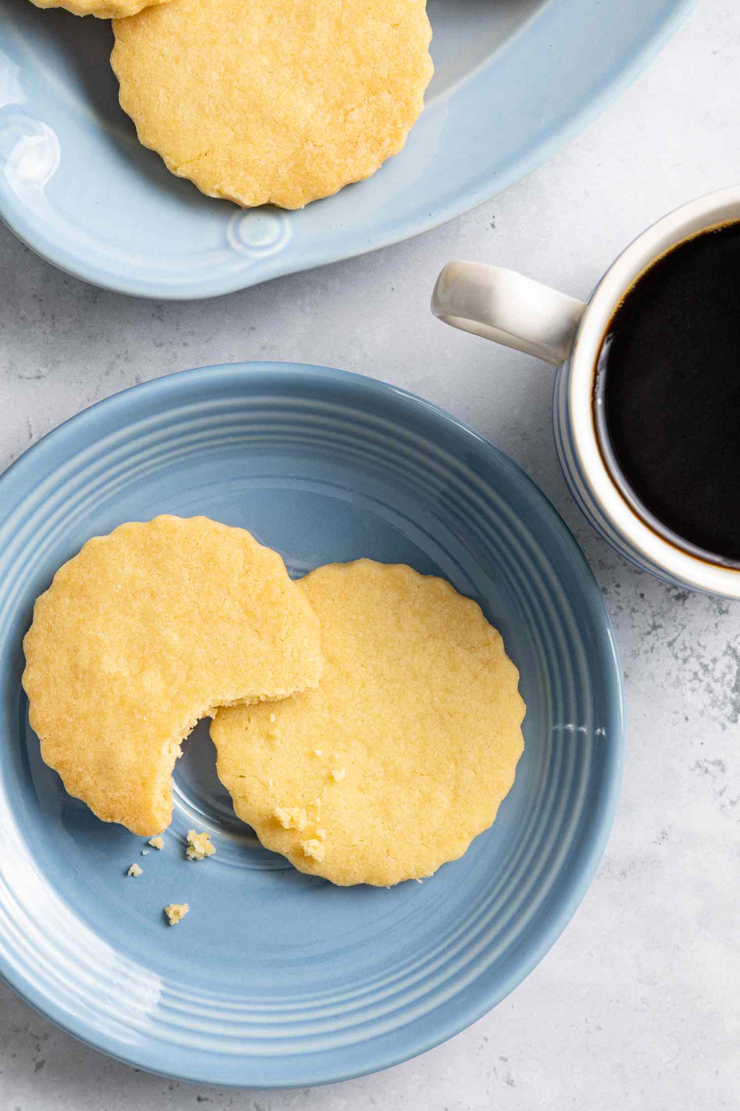 Classic Shortbread Cookies on a Plate with One Bitten from Next to a Plate with More Cookies and a Cup of Coffee