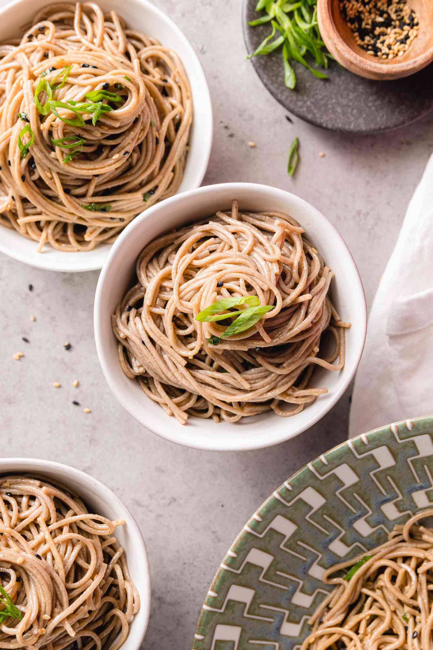 Bowls of Sesame Soba Noodles Recipe Next to a Bigger Bowl with More Noodles and a Plate with Spring Onions and Seasoning