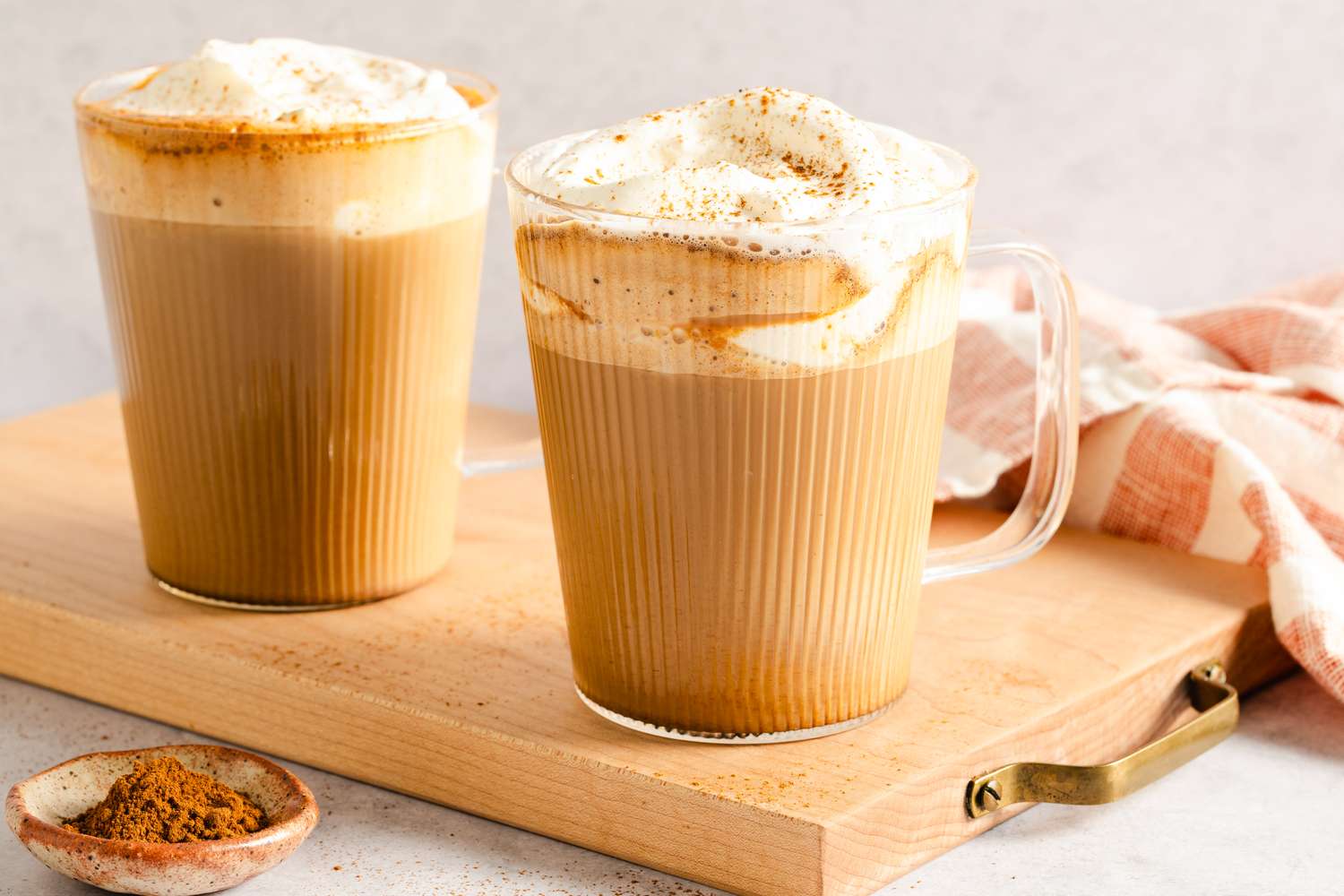 Two frothy pumpkin spice lattes on a wooden board with a small dish of spice nearby