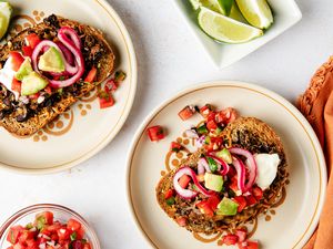 Plates of black bean toast topped with avocado pickled onions and garnishes accompanied by lime wedges and a bowl of salsa