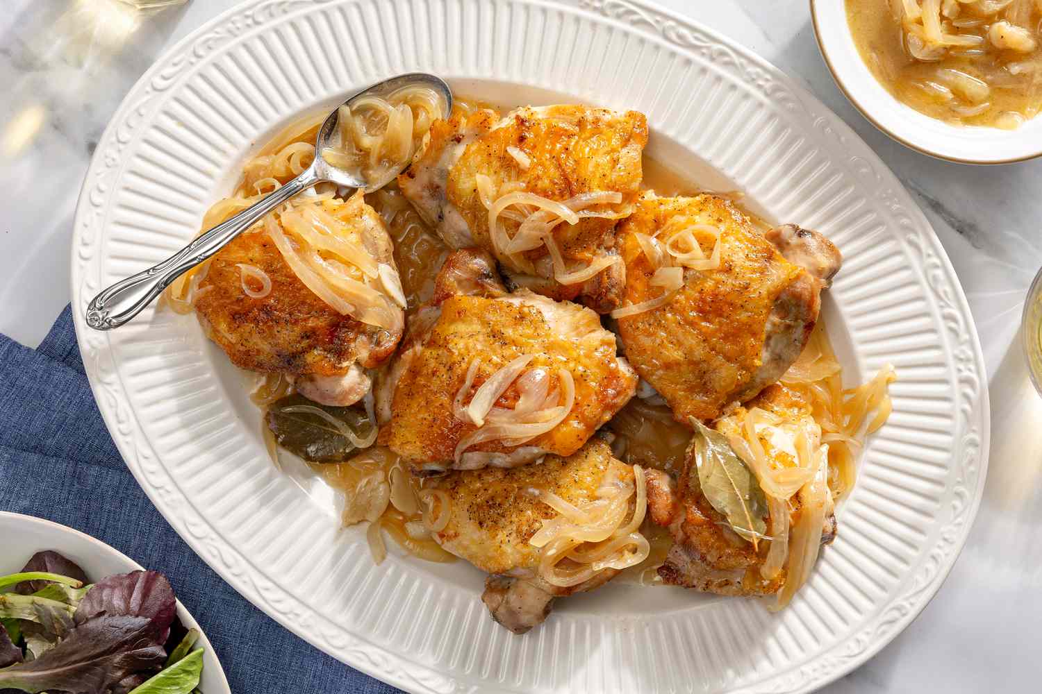Overhead view of a white platter of six wine roasted chicken thighs topped with onions and bay leaves along with a spoon