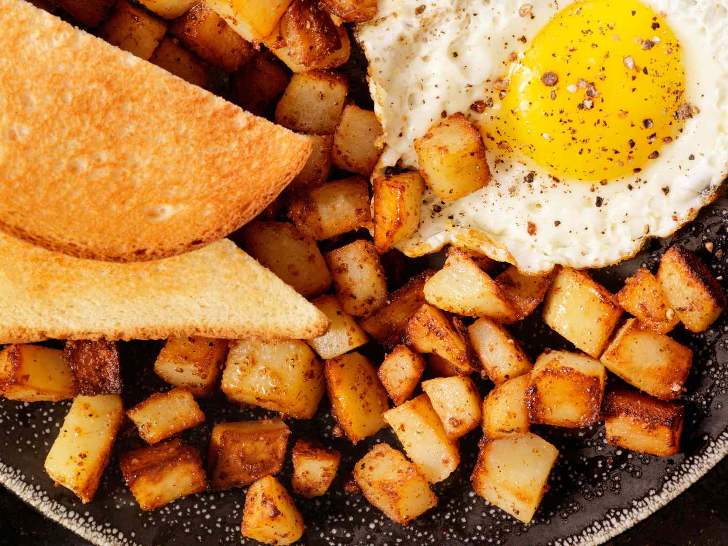 Overhead view of breakfast potatoes on a black plate along with slices of toast and a fried egg
