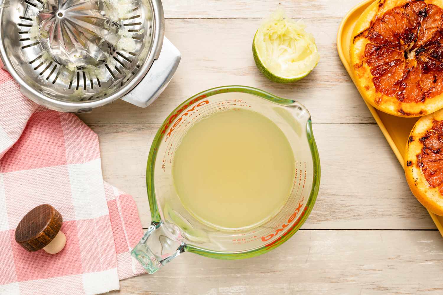 Lime Juice in a Measuring Cup, and in the Surroundings, a Used Lime Half, a Tabletop Juicer, a Tray of Grilled Grapefruit, and a Vodka Bottle Top