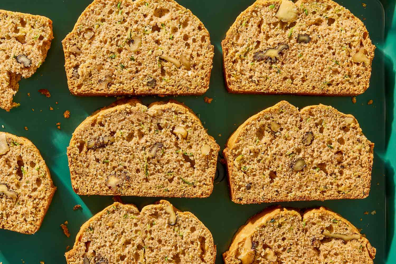 Six slices of zucchini bread arranged on a green surface showcasing nuts and texture