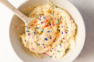 A bowl of yogurt frosting topped with sprinkles, stirred with a spoon