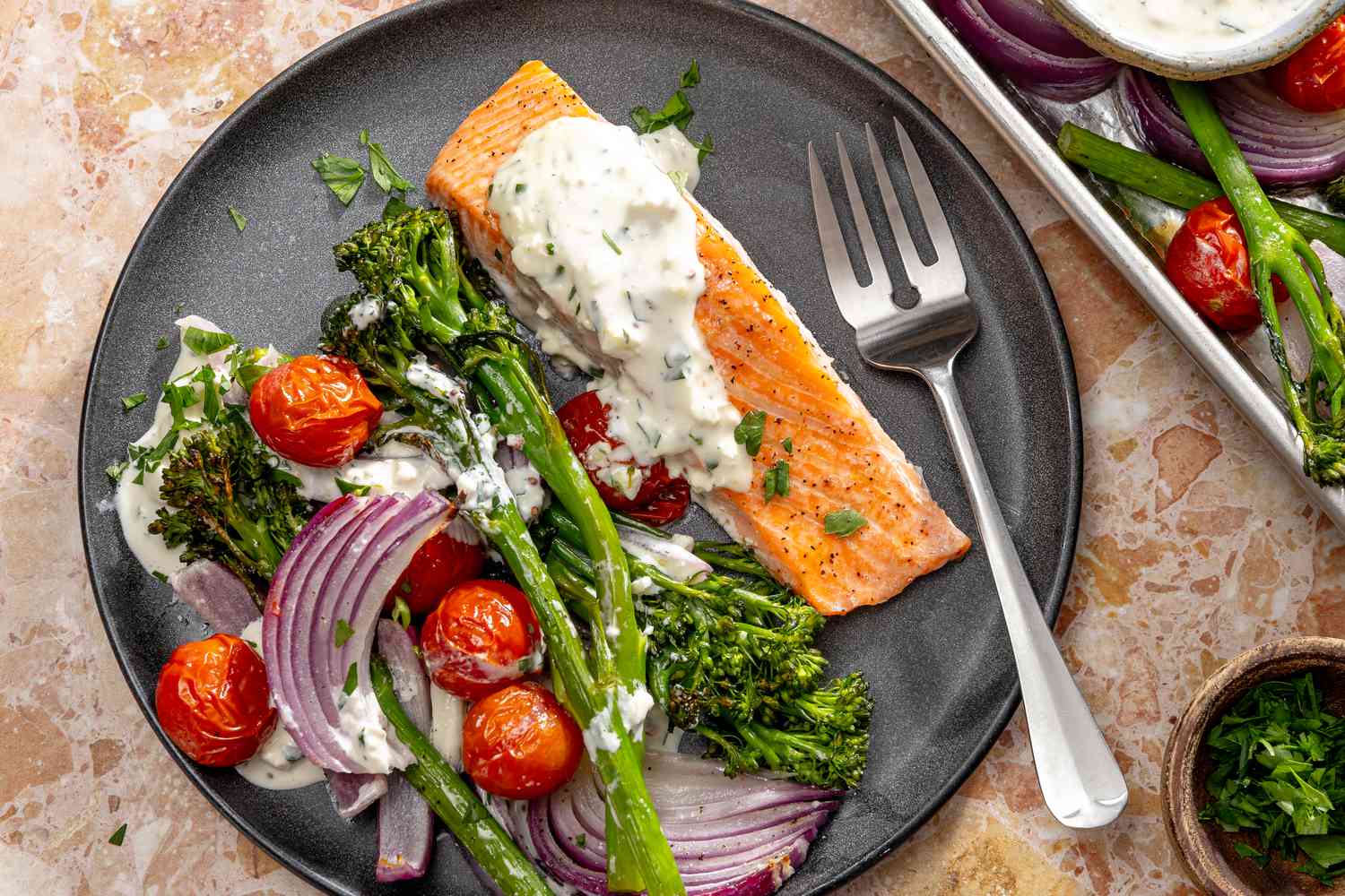 Overhead view of a gray plate of salmon, broccolini cherry tomatoes, onions and feta sauce with a fork on a marble countertop